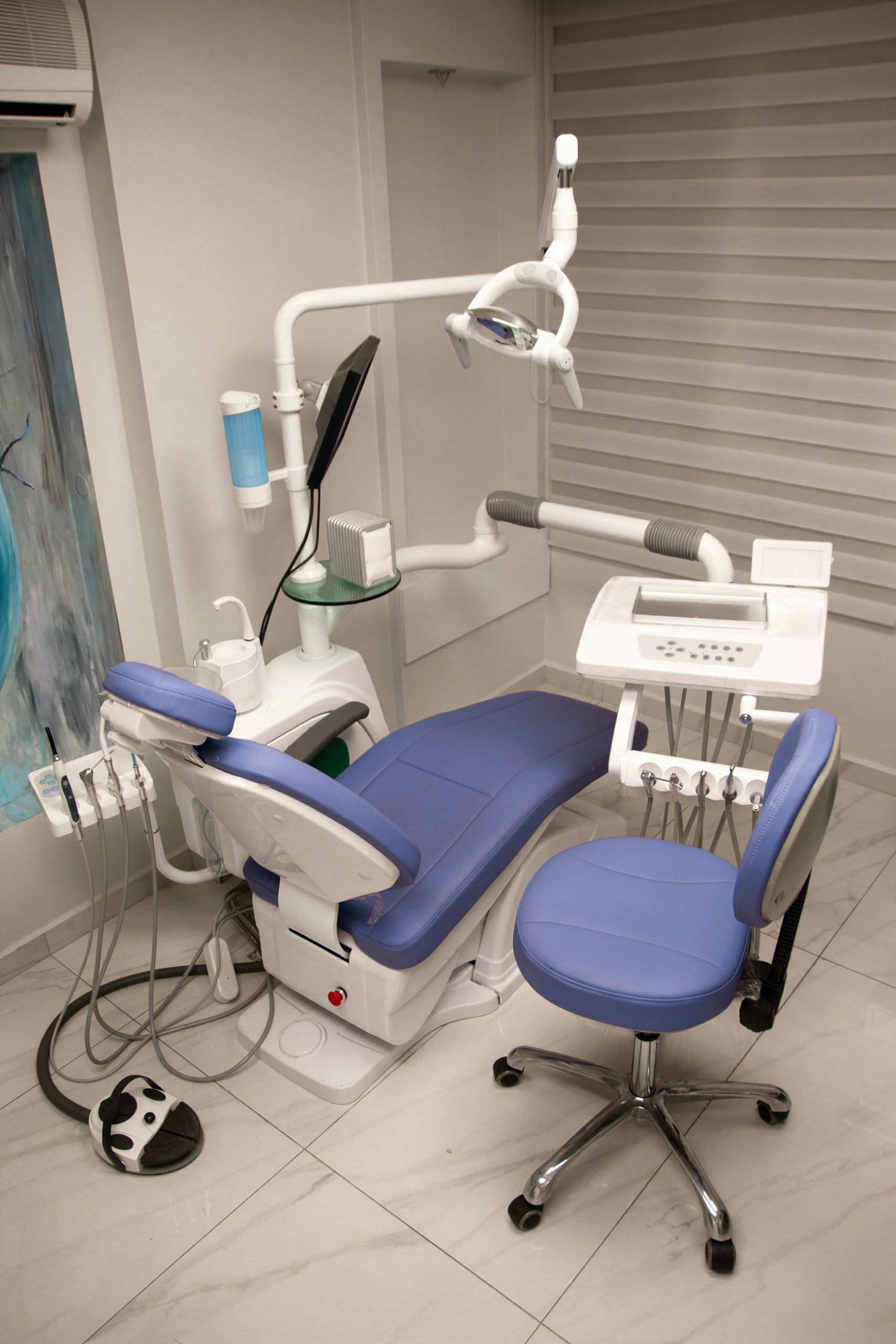 Blue dental chair with overhead lamp, instrument hoses, and control unit in a treatment room.
