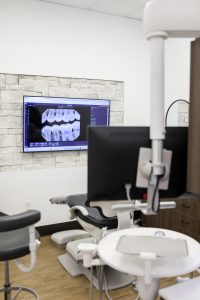 Dental treatment room with chair, monitor, and X-ray displayed on a wall-mounted screen.
