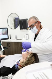 Dentist wearing mask and magnifying loupes treats a patient reclining in a dental chair.