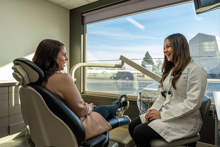Dentist in white coat talks with seated patient in dental chair during consultation.