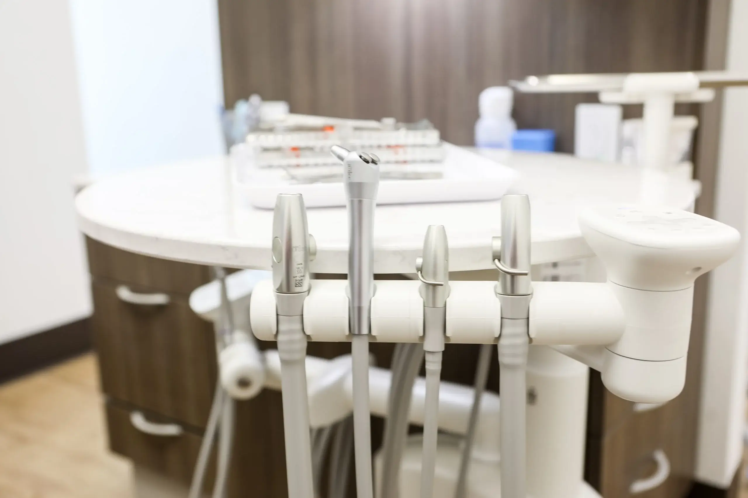 Close-up of dental handpieces and suction hoses on a treatment unit, with instrument tray behind.