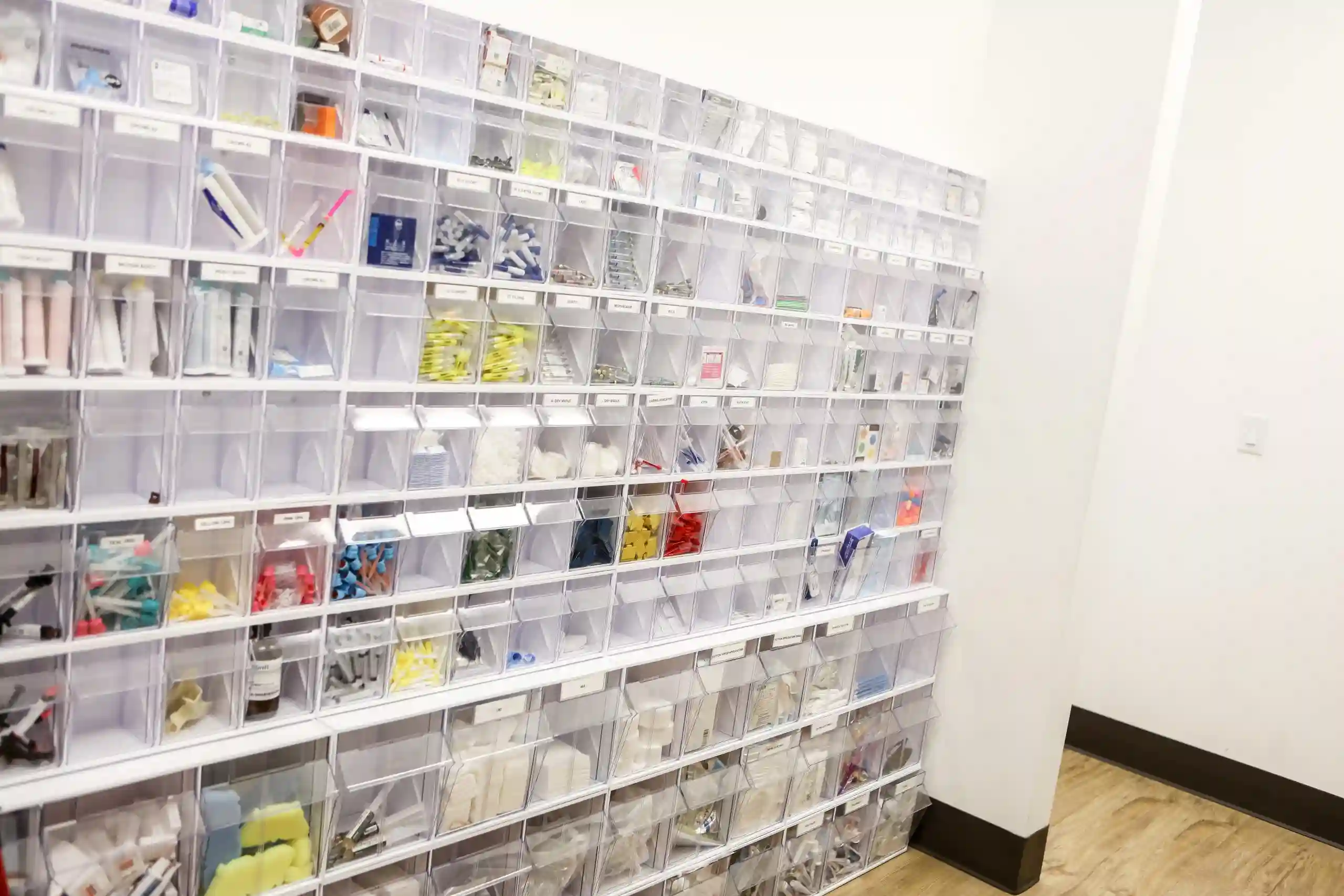Wall of labelled clear storage bins filled with dental supplies in a bright clinic stock room.