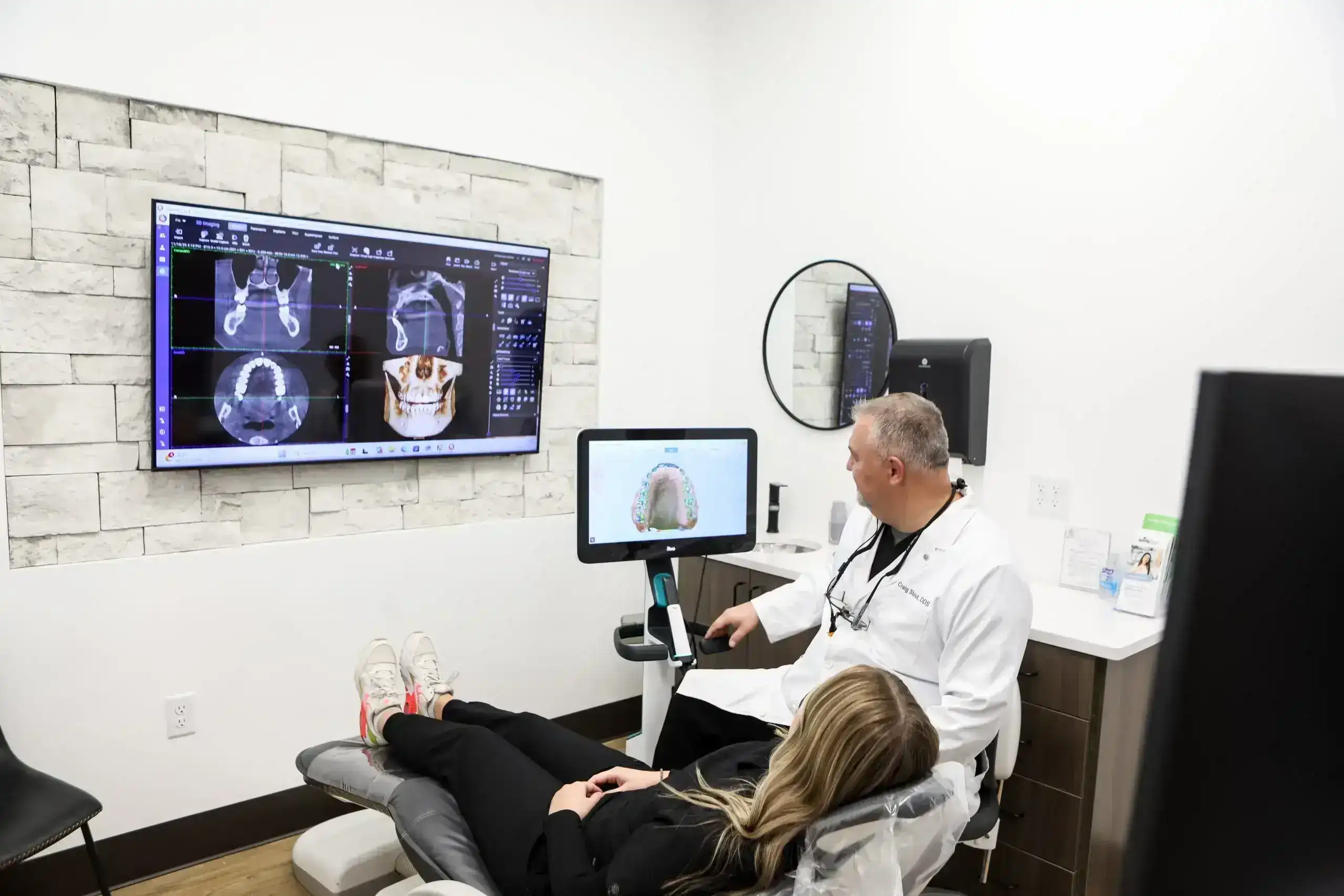 Clinician reviews dental scans on a wall screen while a patient reclines in a treatment chair.