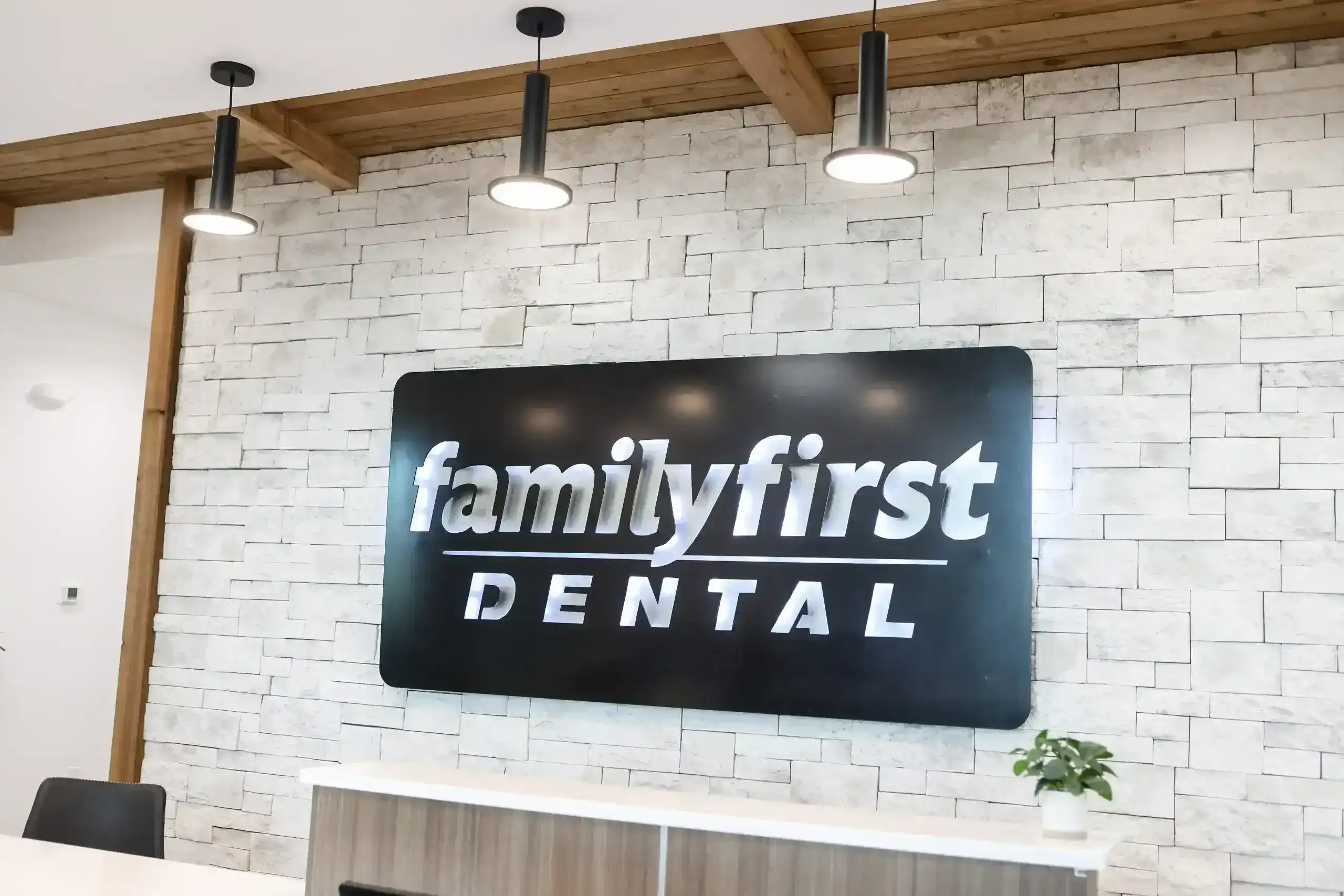 Reception area with a black “familyfirst DENTAL” sign on a white stone wall beneath three hanging lights.
